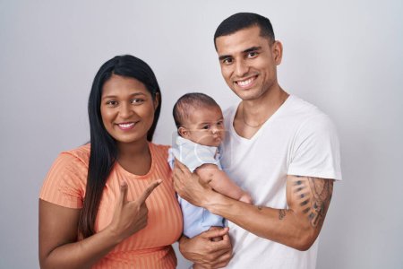 Photo for Young hispanic couple with baby standing together over isolated background cheerful with a smile on face pointing with hand and finger up to the side with happy and natural expression - Royalty Free Image