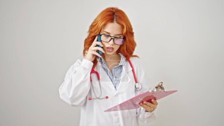 Foto de Mujer pelirroja joven médico leyendo documento en portapapeles hablando en el teléfono inteligente sobre fondo blanco aislado - Imagen libre de derechos