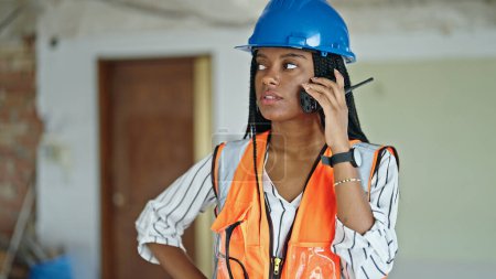 Téléchargez les photos : Femme afro-américaine constructeur debout avec expression détendue parlant par talkie walkie sur le chantier de construction - en image libre de droit