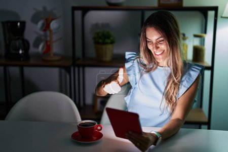 Photo for Young hispanic woman using touchpad sitting on the table at night approving doing positive gesture with hand, thumbs up smiling and happy for success. winner gesture. - Royalty Free Image