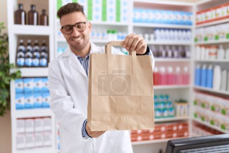 Photo for Young hispanic man pharmacist smiling confident holding shopping bag at pharmacy - Royalty Free Image