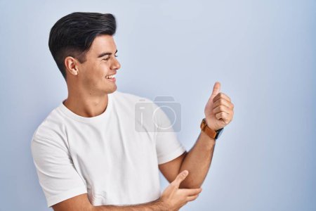 Téléchargez les photos : Homme hispanique debout sur fond bleu regardant fier, souriant faire les pouces geste vers le haut sur le côté - en image libre de droit