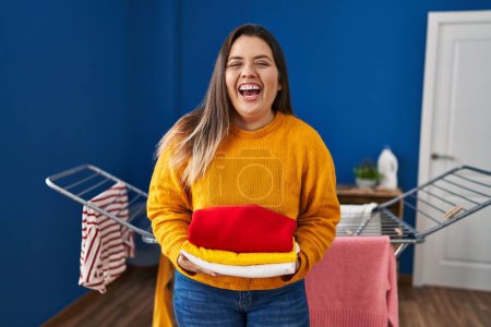 Foto de Mujer hispana joven sosteniendo la ropa limpia y doblada sonriendo y riendo en voz alta porque divertida broma loca. - Imagen libre de derechos