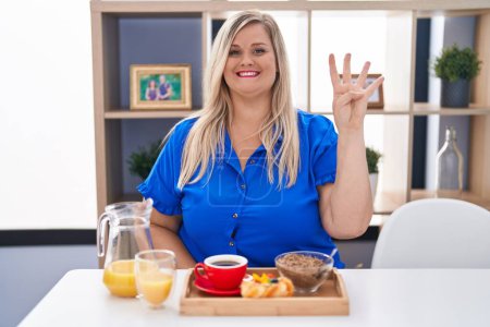 Foto de Caucásico más tamaño mujer desayunando en casa mostrando y apuntando hacia arriba con los dedos número cuatro mientras sonríe confiado y feliz. - Imagen libre de derechos