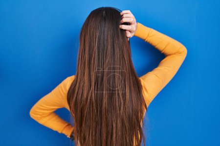 Téléchargez les photos : Jeune femme brune debout sur fond bleu vers l'arrière pensant au doute avec la main sur la tête - en image libre de droit