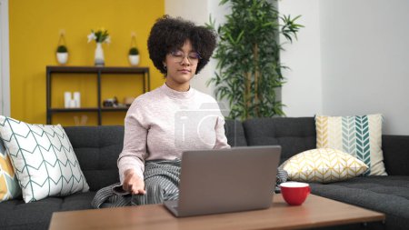 Téléchargez les photos : Jeune femme afro-américaine qui médite assise sur un canapé avec un ordinateur portable à la maison - en image libre de droit