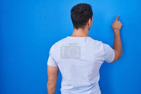 Téléchargez les photos : Jeune homme hispanique debout sur fond bleu posant vers l'arrière pointant vers l'avant avec la main du doigt - en image libre de droit