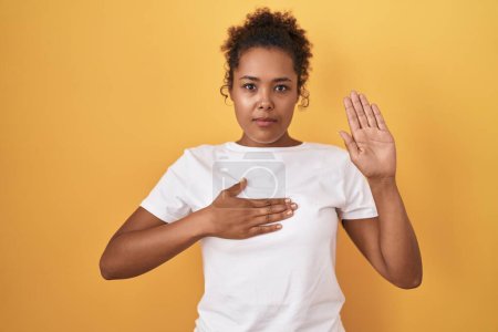 Photo for Young hispanic woman with curly hair standing over yellow background swearing with hand on chest and open palm, making a loyalty promise oath - Royalty Free Image