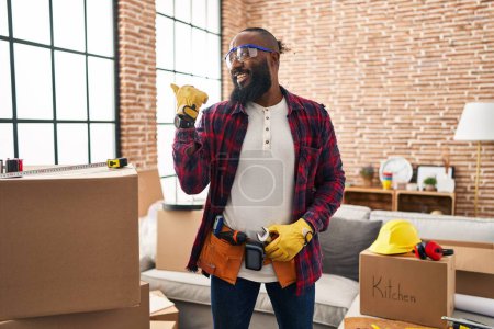 Foto de Hombre afroamericano trabajando en la renovación del hogar sonriendo con la cara feliz mirando y señalando a un lado con el pulgar hacia arriba. - Imagen libre de derechos