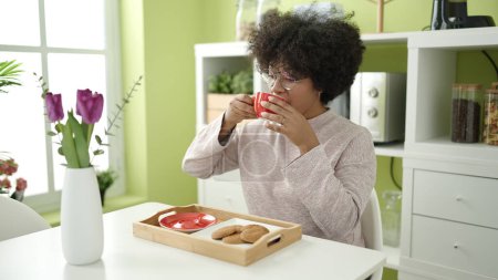 Téléchargez les photos : Jeune femme afro-américaine prenant le petit déjeuner assis sur la table à la maison - en image libre de droit