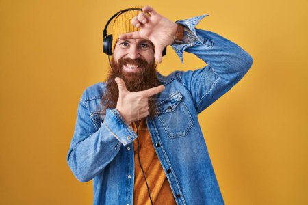Photo for Caucasian man with long beard listening to music using headphones smiling making frame with hands and fingers with happy face. creativity and photography concept. - Royalty Free Image