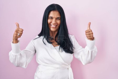 Téléchargez les photos : Femme hispanique mature debout sur fond rose signe de succès faisant geste positif avec la main, pouces levés souriant et heureux. expression joyeuse et geste gagnant. - en image libre de droit