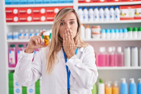 Foto de Mujer rubia joven que trabaja en la farmacia sosteniendo píldoras que cubren la boca con la mano, conmocionada y temerosa de equivocarse. expresión sorprendida - Imagen libre de derechos