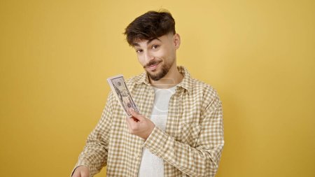 Téléchargez les photos : Jeune homme arabe souriant confiant tenant des dollars sur fond jaune isolé - en image libre de droit