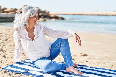 Foto de Middle age woman smiling confident sitting on towel at seaside - Imagen libre de derechos