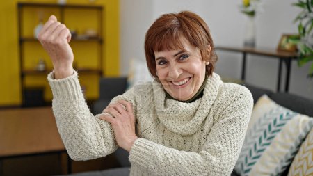 Foto de Mujer hispana madura sonriendo confiada haciendo un gesto fuerte con el brazo en casa - Imagen libre de derechos