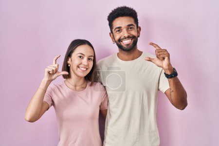 Téléchargez les photos : Jeune couple hispanique ensemble sur fond rose geste souriant et confiant avec la main faisant signe de petite taille avec les doigts à la recherche et la caméra. concept de mesure. - en image libre de droit