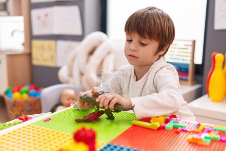 Foto de Adorable chico caucásico jugando con dinosaurio juguete sentado en la mesa en el jardín de infantes - Imagen libre de derechos