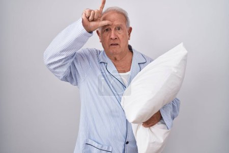 Photo for Senior man with grey hair wearing pijama hugging pillow making fun of people with fingers on forehead doing loser gesture mocking and insulting. - Royalty Free Image