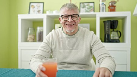 Téléchargez les photos : Moyen-âge homme aux cheveux gris boire un verre de jus assis sur la table à la maison - en image libre de droit