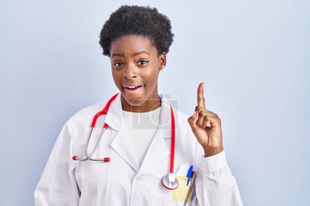Téléchargez les photos : Femme afro-américaine portant un uniforme de médecin et un stéthoscope pointant du doigt avec une idée réussie. Sorti et heureux. numéro un. - en image libre de droit