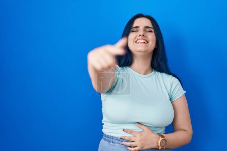 Photo for Young modern girl with blue hair standing over blue background laughing at you, pointing finger to the camera with hand over body, shame expression - Royalty Free Image