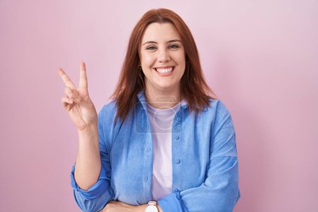 Foto de Mujer hispana joven con el pelo rojo de pie sobre fondo rosa sonriendo con la cara feliz guiñando el ojo a la cámara haciendo signo de victoria con los dedos. número dos. - Imagen libre de derechos