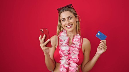 Foto de Mujer rubia joven que usa bikini de compras con teléfono inteligente y tarjeta de crédito sobre fondo rojo aislado - Imagen libre de derechos