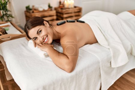 Photo for Young beautiful hispanic woman lying on table having hot stones massage at beauty salon - Royalty Free Image