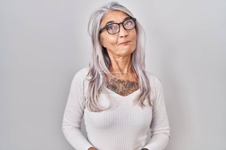Foto de Mujer de mediana edad con el pelo gris de pie sobre fondo blanco sonriendo mirando a un lado y mirando hacia otro pensando. - Imagen libre de derechos