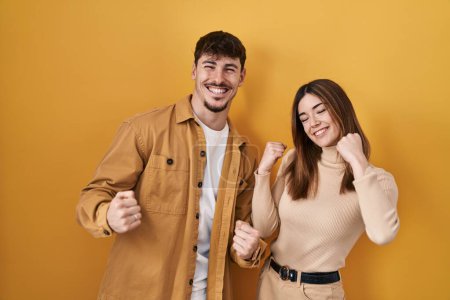 Photo for Young hispanic couple standing over yellow background very happy and excited doing winner gesture with arms raised, smiling and screaming for success. celebration concept. - Royalty Free Image