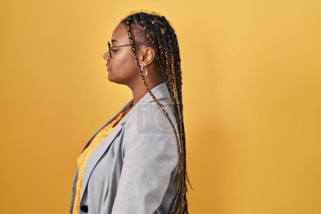 Téléchargez les photos : Femme afro-américaine avec des tresses debout sur fond jaune regardant vers le côté, pose de profil relax avec visage naturel avec sourire confiant. - en image libre de droit