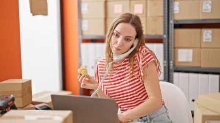 Photo for Young blonde woman ecommerce business worker eating croissant talking on telephone working at office - Royalty Free Image