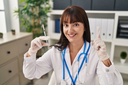 Téléchargez les photos : Jeune femme brune médecin tenant une seringue surprise avec une idée ou une question pointant du doigt avec un visage heureux, numéro un - en image libre de droit