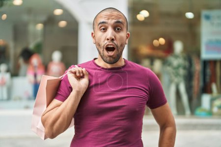 Photo for Hispanic man with beard holding shopping bags outdoors scared and amazed with open mouth for surprise, disbelief face - Royalty Free Image