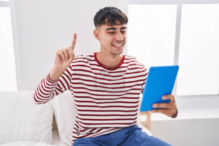 Téléchargez les photos : Jeune homme hispanique utilisant pavé tactile assis sur le lit souriant avec une idée ou une question pointant du doigt avec le visage heureux, numéro un - en image libre de droit