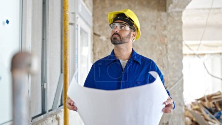 Photo for Young hispanic man worker wearing hardhat reading house project at construction site - Royalty Free Image