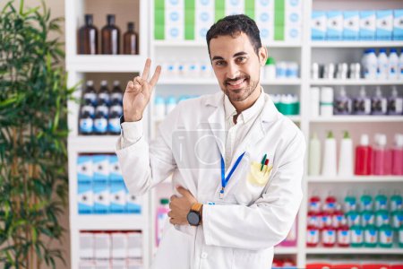 Foto de Hombre hispano guapo trabajando en farmacia sonriendo mirando a la cámara mostrando los dedos haciendo señal de victoria. número dos. - Imagen libre de derechos