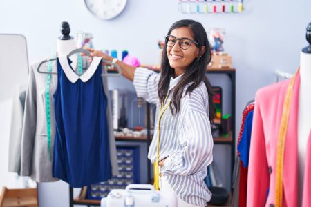 Téléchargez les photos : Jeune belle femme hispanique tailleur souriant confiant tenant t-shirt à l'usine de vêtements - en image libre de droit