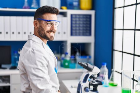 Foto de Joven hombre hispano vistiendo uniforme científico sonriendo confiado en laboratorio - Imagen libre de derechos