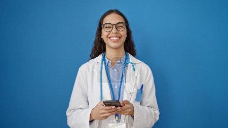 Foto de Joven hermosa mujer hispana médico sonriendo con teléfono inteligente sobre fondo azul aislado - Imagen libre de derechos