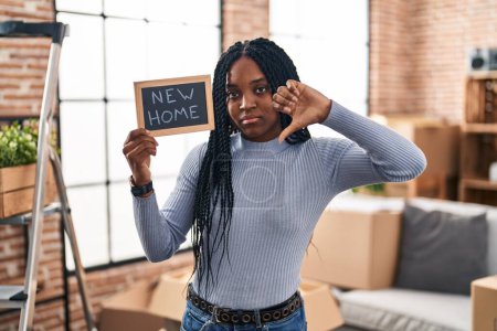 Téléchargez les photos : Femme afro-américaine tenant tableau noir avec nouveau texte à la maison avec le visage en colère, signe négatif montrant aversion avec les pouces vers le bas, concept de rejet - en image libre de droit