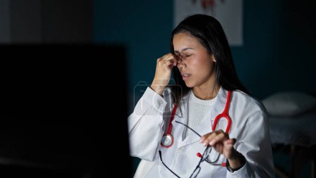 Foto de Joven hermosa mujer hispana médico estresado trabajando sacando gafas en la clínica - Imagen libre de derechos