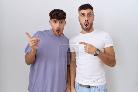 Foto de Homosexual pareja gay de pie sobre blanco fondo sorprendido señalando con el dedo a un lado, boca abierta asombrado expresión. - Imagen libre de derechos