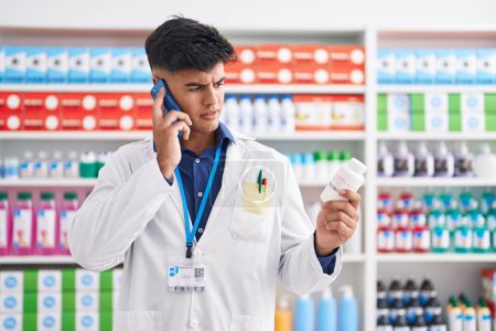 Téléchargez les photos : Jeune homme hispanique pharmacien tenant bouteille de pilules parler sur smartphone à la pharmacie - en image libre de droit