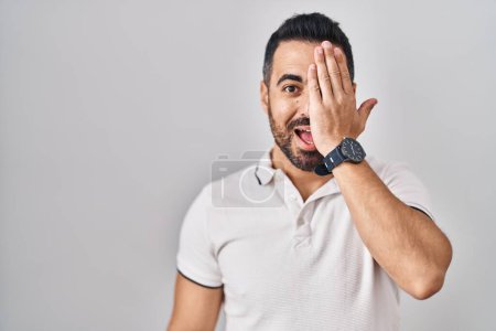 Photo for Young hispanic man with beard wearing casual clothes over white background covering one eye with hand, confident smile on face and surprise emotion. - Royalty Free Image