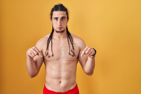 Photo for Hispanic man with long hair standing shirtless over yellow background pointing down looking sad and upset, indicating direction with fingers, unhappy and depressed. - Royalty Free Image