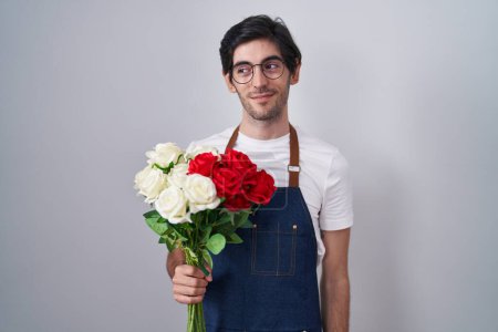 Foto de Joven hombre hispano sosteniendo un ramo de rosas blancas y rojas sonriendo mirando hacia un lado y mirando hacia otro pensando. - Imagen libre de derechos