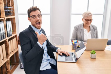 Téléchargez les photos : Jeune homme hispanique travaillant au bureau pointant le pouce vers le côté souriant heureux avec la bouche ouverte - en image libre de droit
