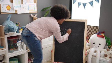Téléchargez les photos : Jeune femme afro-américaine prof de maternelle écrivant sur tableau noir à la maternelle - en image libre de droit
